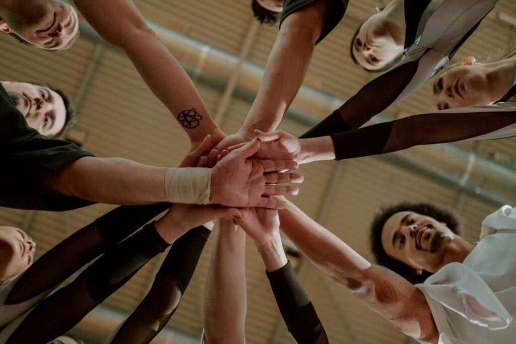 Close-up of multiple hands stacked together in a team gesture. The photo, taken from below, shows smiling men and women, conveying collaboration, unity, and teamwork.