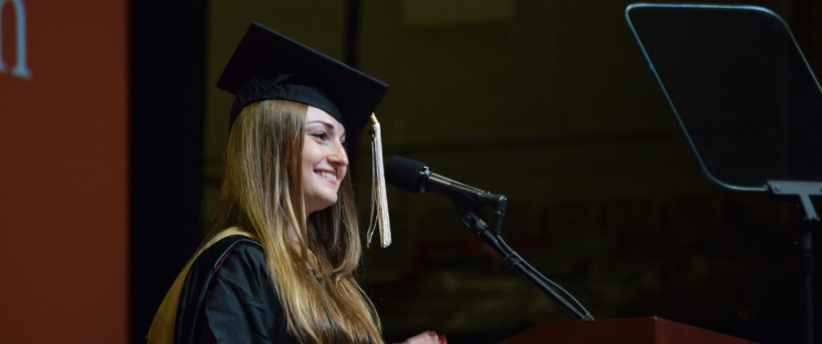 Photo of a woman wearing a graduation cap and gown, she is looking out into a crowd and facing a microphone