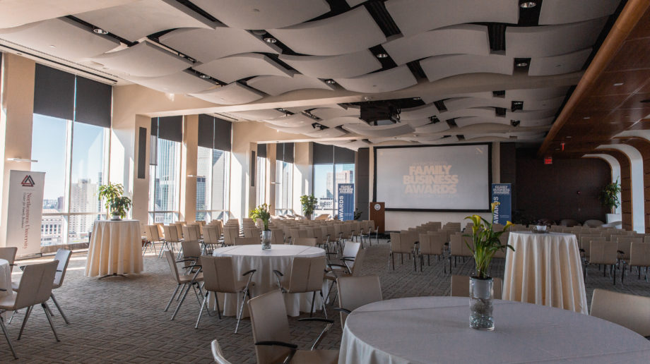Photograph of a large room with tables and chairs set up for a reception and a screen in the front of the room reading 'Family Business Awards'