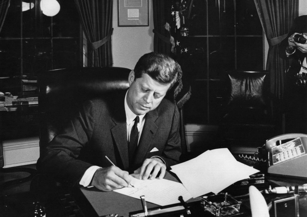 Black and white photograph of President John F. Kennedy sitting in his office signing a document