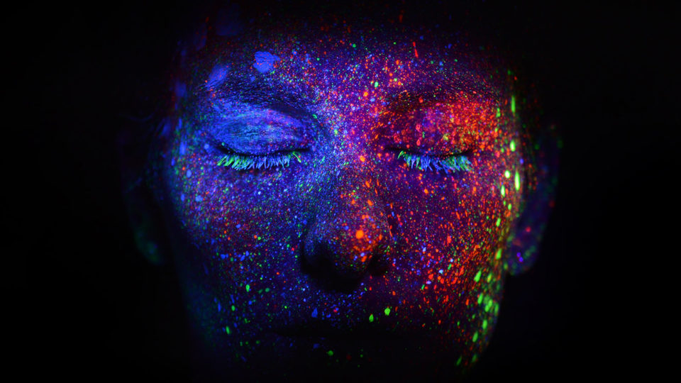 Photograph of a persons face covered in colorful splotches of glow in the dark paint