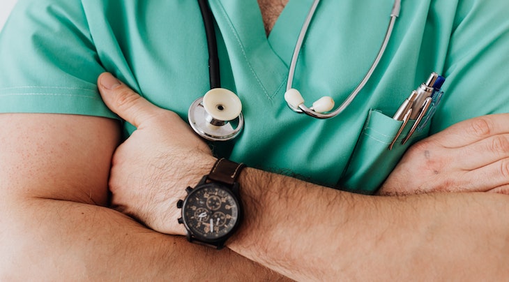 Photo of a man with his arms crossed in front of him wearing a watch, scrubs, a stethoscope, and pens in his shirt pocket