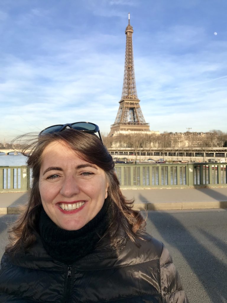 photo of a woman with brown hair and sunglasses standing in front of the Eiffel tower