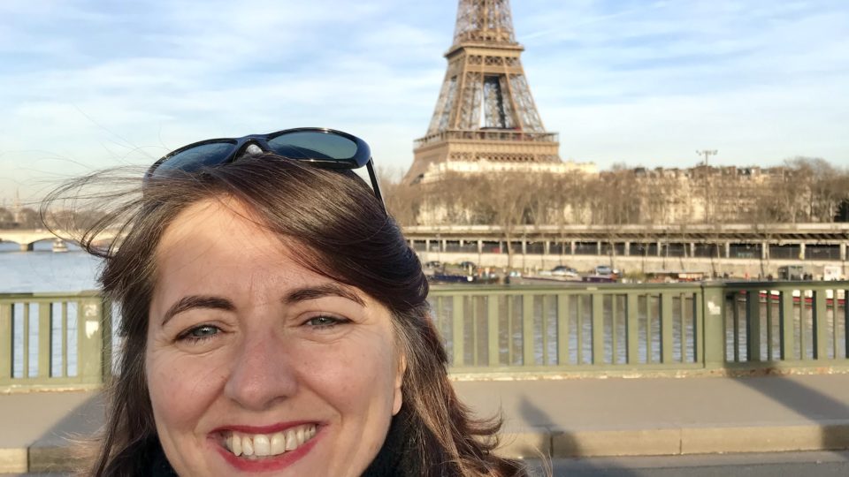 photo of a woman with brown hair and sunglasses standing in front of the Eiffel tower