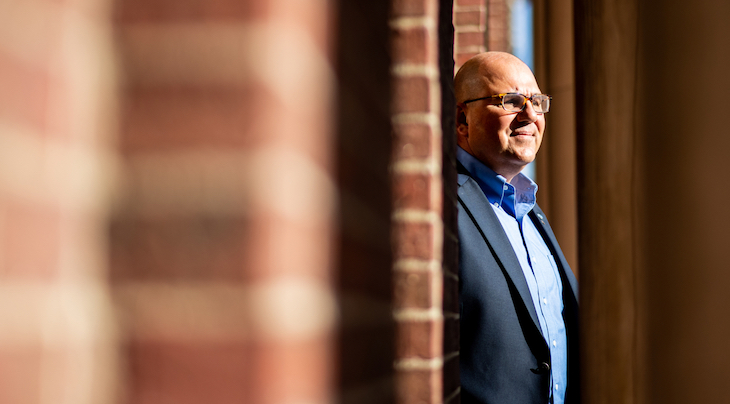 Photo of Chris Dewitt, man wearing a blue button down with a blazer, wearing glasses and looking into the sun while leaning against a brick building