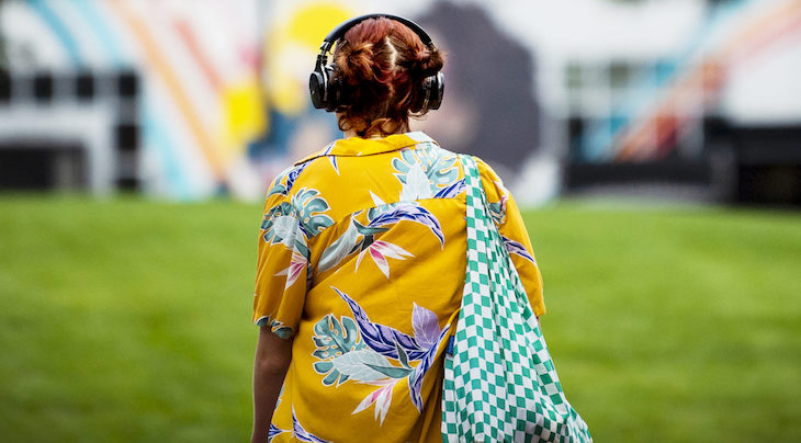 Photo of a person wearing a yellow floral collared shirt and a green checker print bag with black headphones walking away from the camera