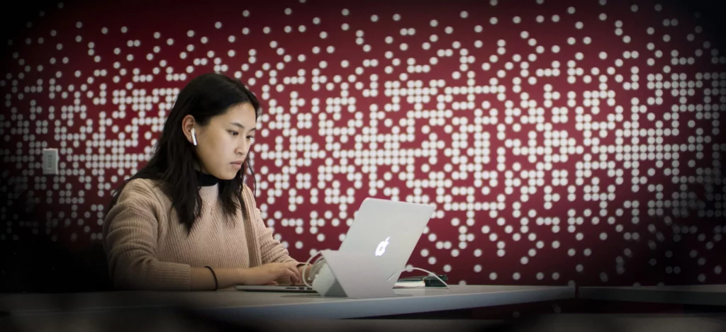 photo of a female student with long dark hair wearing airpods and sitting at a table typing on a laptop