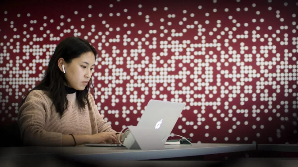 photo of a female student with long dark hair wearing airpods and sitting at a table typing on a laptop