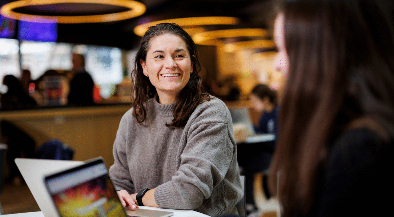 A smiling young woman with dark hair, wearing a gray turtleneck sweater and a fitness tracker, sits at a table with an open laptop in a casual indoor campus common area. She is engaged in conversation with another person seated across from her, whose back is partially visible in the foreground. The background shows a warmly lit space with other people and glowing circular ceiling lights.