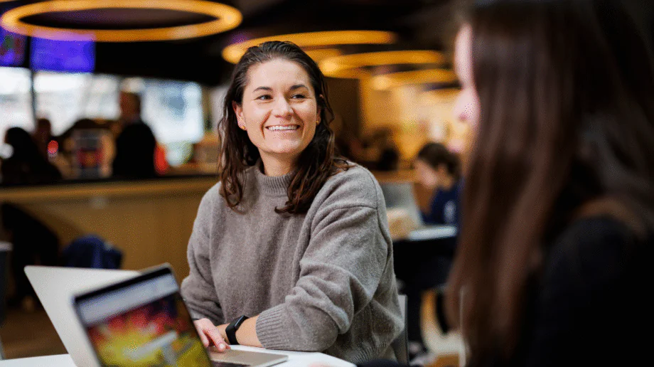A smiling young woman with dark hair, wearing a gray turtleneck sweater and a fitness tracker, sits at a table with an open laptop in a casual indoor campus common area. She is engaged in conversation with another person seated across from her, whose back is partially visible in the foreground. The background shows a warmly lit space with other people and glowing circular ceiling lights.