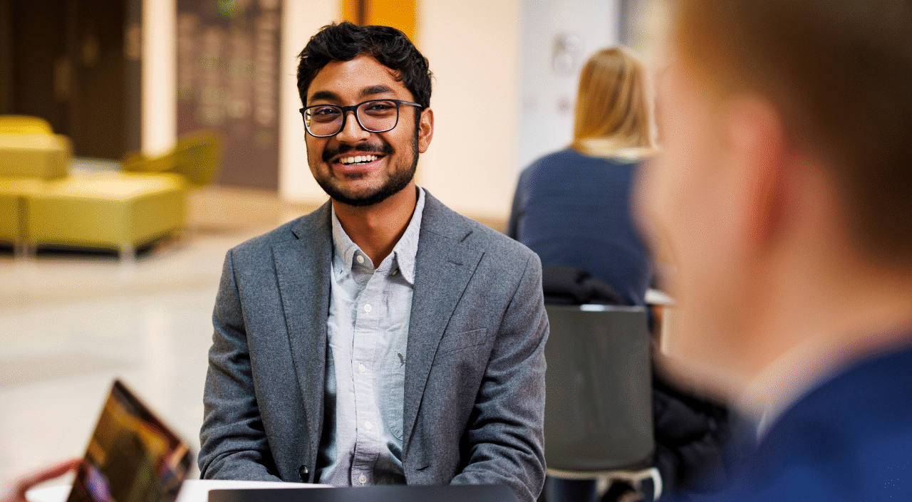 A young man with glasses and a beard smiles warmly while seated at a table in a university common area, appearing engaged in conversation. He wears a gray blazer over a light blue shirt. In the background, a woman with blonde hair sits at another table, and yellow seating is visible in the lounge area behind them.