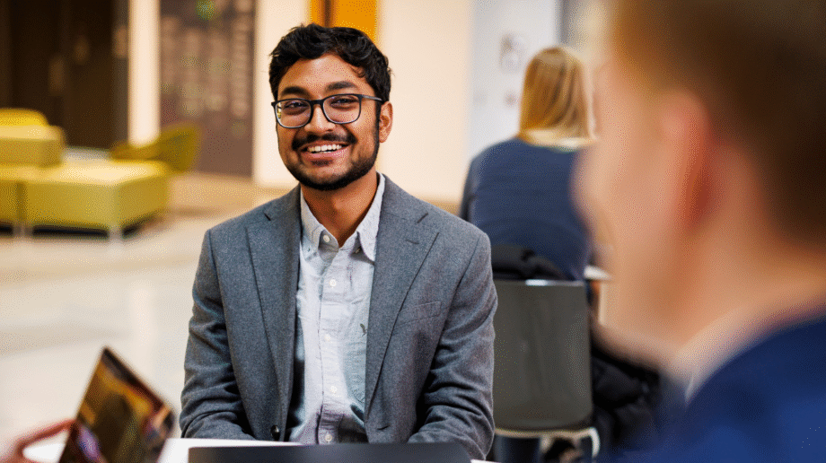 A young man with glasses and a beard smiles warmly while seated at a table in a university common area, appearing engaged in conversation. He wears a gray blazer over a light blue shirt. In the background, a woman with blonde hair sits at another table, and yellow seating is visible in the lounge area behind them.