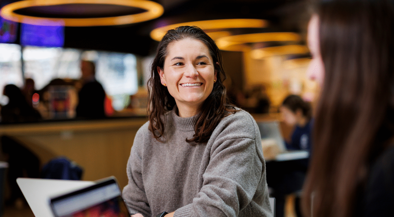 A smiling woman with dark hair wearing a gray turtleneck sweater sits in a modern café or lounge, looking toward someone across from her. A laptop is partially visible in the foreground. Other people working on laptops are blurred in the background, with warm overhead ring lighting.