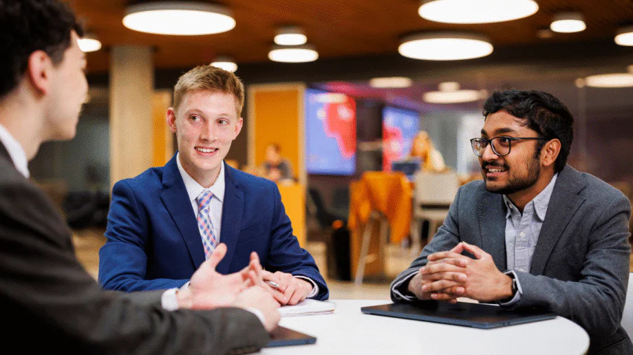 Three professionally dressed young men having a conversation at a round white table in a modern, well-lit café or university common area. Two men face the camera — one wearing a blue suit with a pink plaid tie, the other in a gray blazer and glasses — while a third person with their back to the camera gestures as they speak. Laptops and notebooks are on the table. In the background, other people work at tables near orange partition walls and a large display screen.