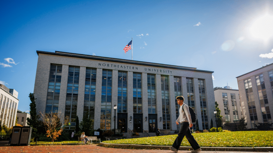 A student with a backpack walks past a Northeastern University academic building on a sunny day, with an American flag flying above the entrance and green lawns in the foreground.